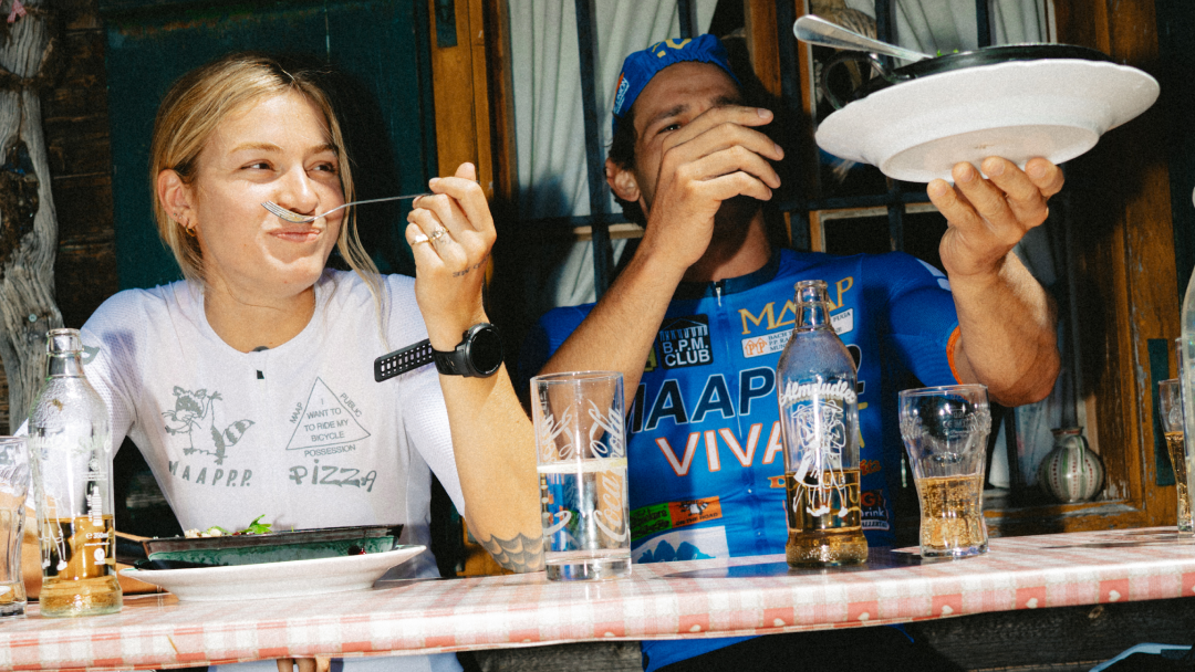 Two people sitting at a rustic wooden table in a restaurant, eating and drinking. The person on the left wears a white graphic T-shirt, black shorts, white socks, and white sneakers.