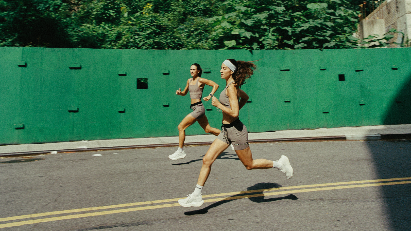 Two women running on a city street wearing athletic apparel, with a green construction fence, trees, and urban buildings in the background.