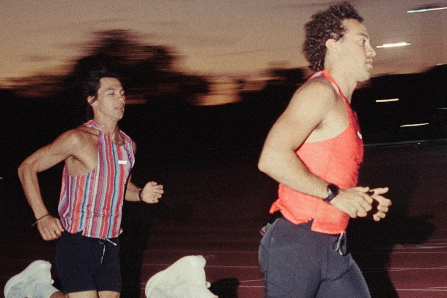 Two men running on an athletic track at dusk, one wearing a red, white, and blue striped tank top and black shorts, and the other wearing a bright coral tank top and dark shorts.