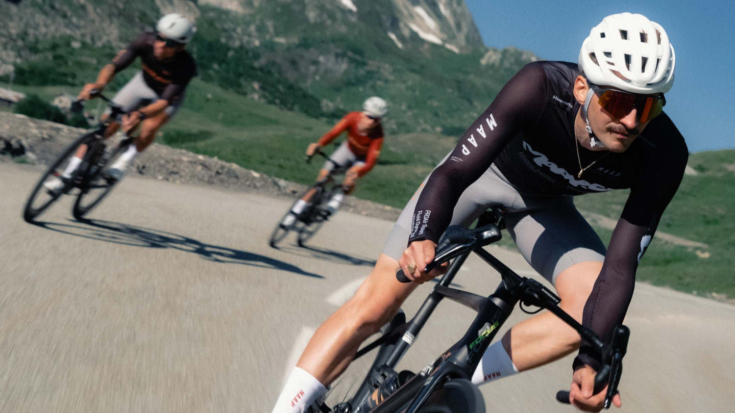 A cyclist in a black and gray cycling jersey rides a road bike on a winding mountain road, with other cyclists and green hills in the background. MAAP