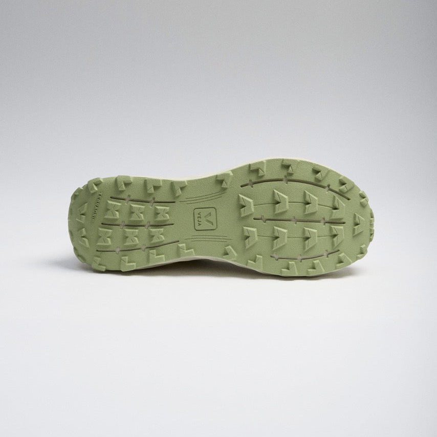 The outsole of a green shoe, showing a textured tread pattern and a small embossed logo, photographed against a neutral gray studio background.