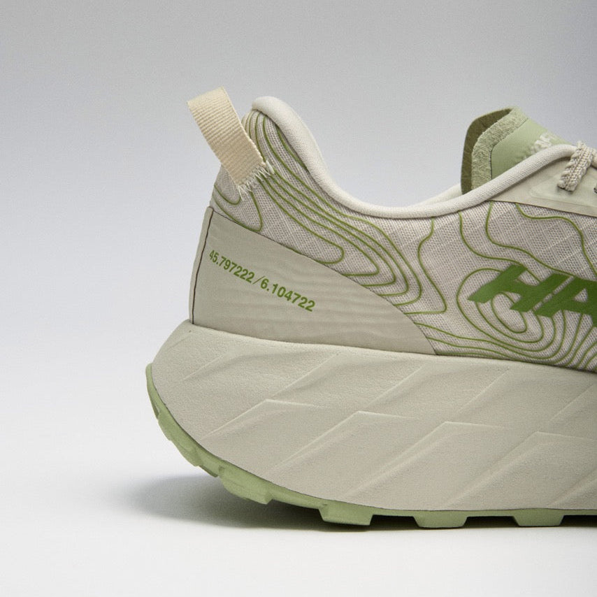 Close-up side view of a Hoka sneaker in off-white and light green, showing the heel, midsole, and part of the upper with green contour-style line graphics and branding.